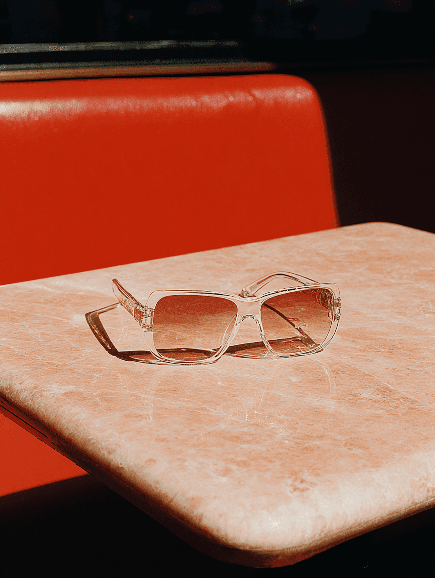 Clear sunglasses resting on a pink marble table in front of a red booth
